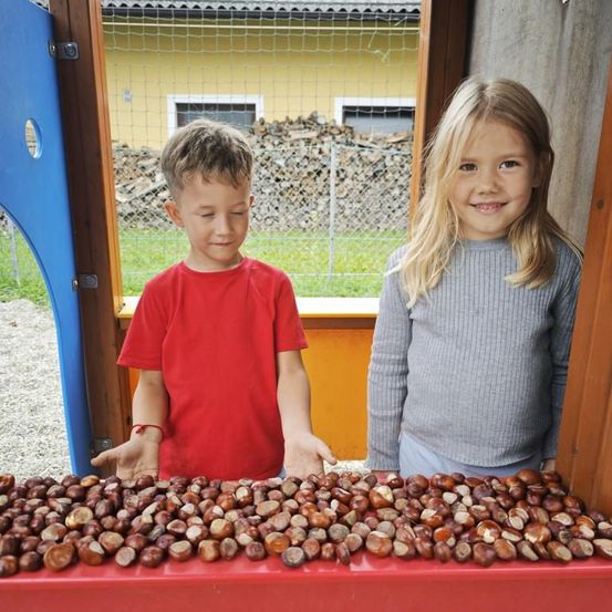 Zwei Kinder stehen hinter einem Tisch voller Kastanien, mit einem gelben Haus und einem eingezäunten Hof im Hintergrund.