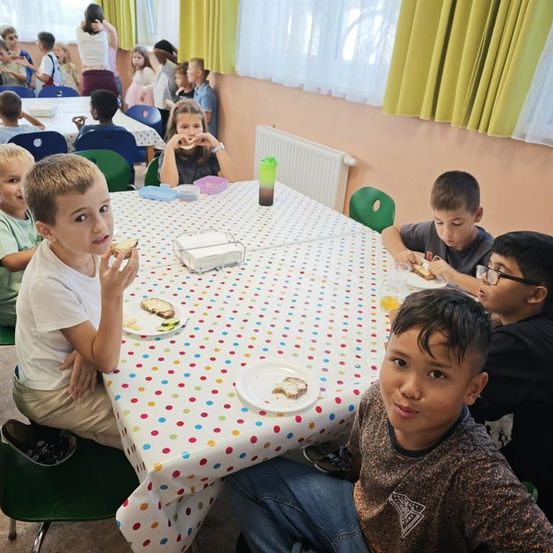 Mehrere Kinder sitzen um einen Tisch und essen Sandwiches, ein Junge lächelt in die Kamera. Der Tisch hat eine Tischdecke mit Punkten.