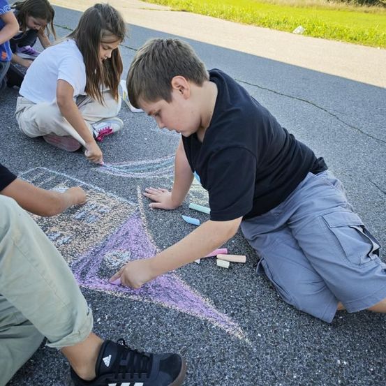 Mehrere Kinder sind am Boden und zeichnen mit Kreide. Ein Junge in Schwarz arbeitet an einem Stern. Neben ihm ist ein Mädchen in Weiß.