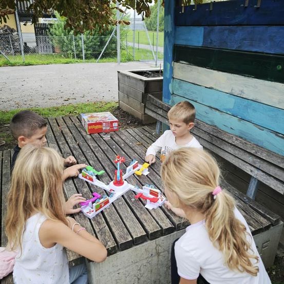 Vier Kinder spielen draußen am Tisch ein Brettspiel. Sie sitzen auf einer Holzbank. Dahinter befindet sich eine farbenfrohe Wand mit einem Topf.