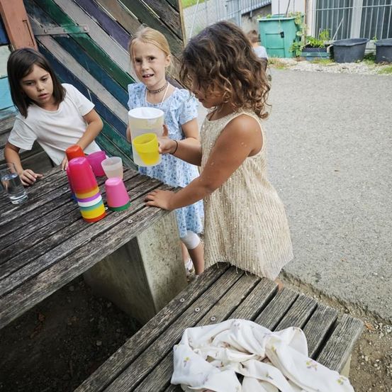 Drei Mädchen spielen mit bunten Bechern auf einer Picknickbank, eines hält einen gelben Becher, ein anderes schaut auf ein Glas Wasser. Eine Wand mit leuchtenden Farben und grünen Behältern ist dahinter.