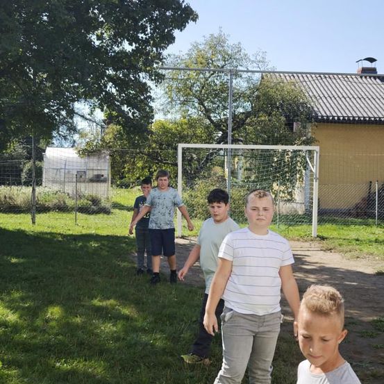 Vier Jungen spielen auf einem Feld mit einem Fußballtor im Hintergrund. Sie gehen auf die Kamera zu.