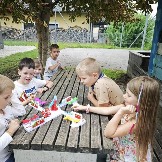 Eine Gruppe von Kindern spielt mit einem Spielzeug auf einem Holztisch in einem Park. Der Junge in der Mitte lehnt sich vor und das Mädchen neben ihm schaut ihn an. Dahinter steht ein Baum mit einem Zaun und einem Haus.