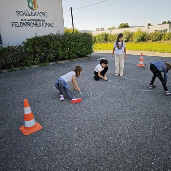 Mehrere junge Mädchen spielen auf einem gepflasterten Grundstück mit orangefarbenen Verkehrskegel. Sie zeichnen auf den Boden. Ein Gebäude mit dem Schild 'Schulerhort der Marktgemeinde Feldkirchen/Graz' steht dahinter.