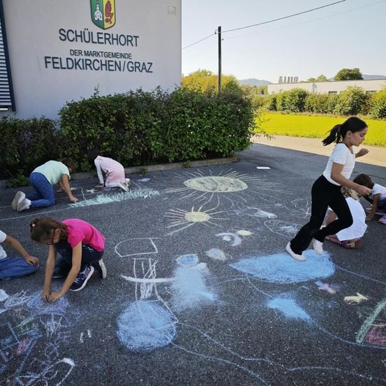 Kinder zeichnen mit Kreide auf dem Boden vor einem Gebäude mit der Aufschrift Schulerhort in Feldkirchen/Graz.