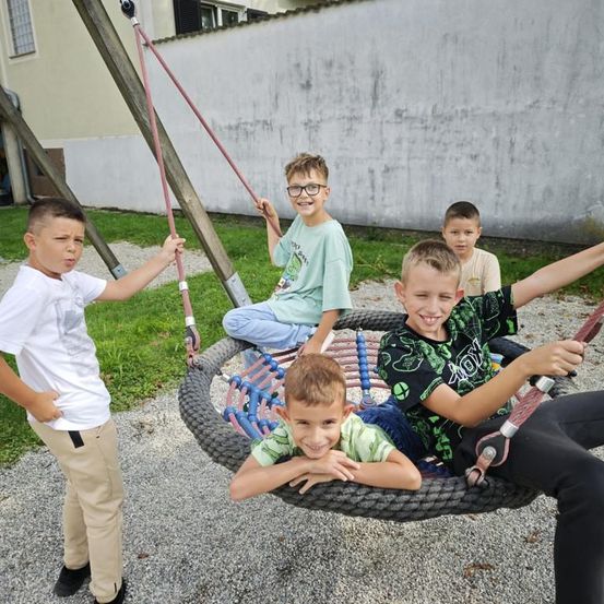 Fünf Jungen sind auf einem Spielplatz. Ein Junge sitzt auf einem Reifenschaukel, ein anderer Junge steht neben ihm und drei Jungen lächeln. Dahinter ist eine weiße Wand.