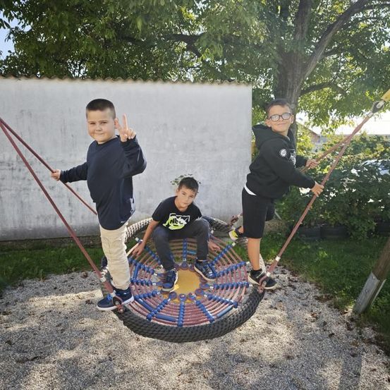 Drei junge Jungen genießen eine Schaukel im Park, einer macht ein Friedenszeichen. Sie sind von Kies und Grün umgeben.