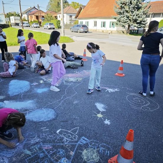 Eine Gruppe von Kindern zeichnet mit Kreide auf der Straße. Mehrere Autos sind am Straßenrand geparkt.