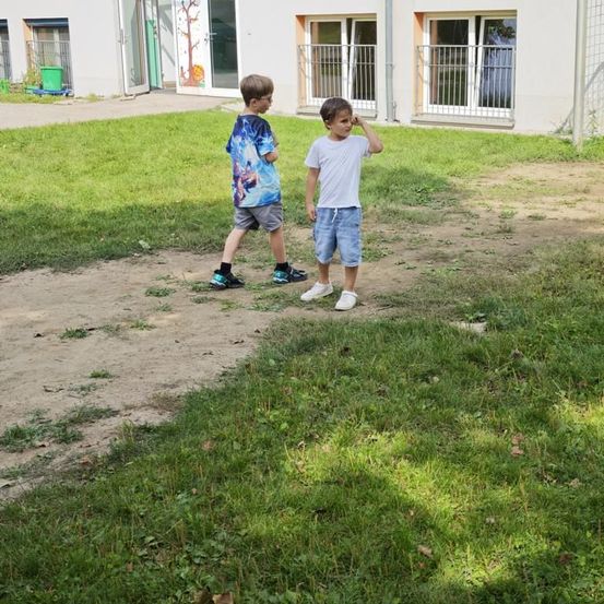 Zwei junge Jungen gehen auf einem Außenbereich. Ein Junge trägt ein blaues T-Shirt und Shorts, der andere trägt ein weißes T-Shirt und Shorts. Der Bereich ist von Gras umgeben.