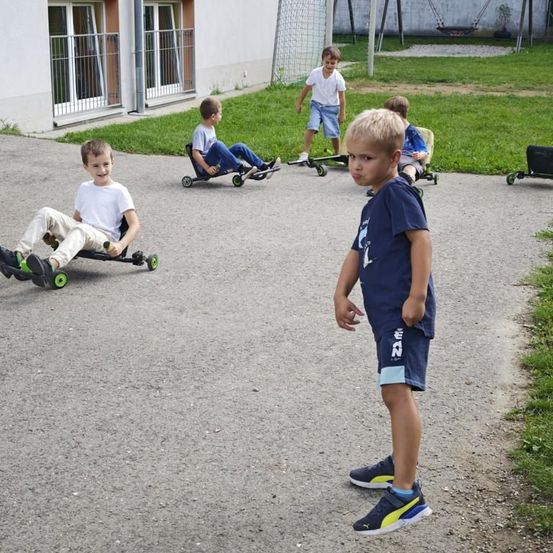 Fünf junge Jungen fahren auf kleinen Rädern in einem Außenbereich. Ein Junge steht und beobachtet. Der Boden ist asphaltiert, mit Gras und einem Gebäude im Hintergrund.