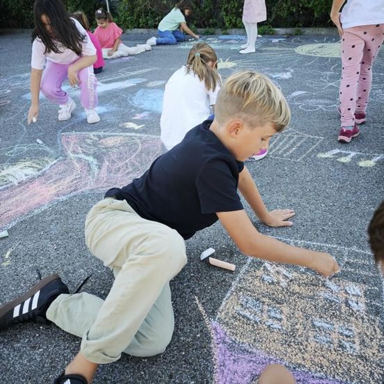 Mehrere Kinder sind auf einem Bürgersteig und zeichnen mit Kreide auf den Boden. Ein Junge kniet und konzentriert sich auf seine Zeichnung.