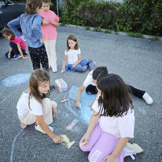 Mehrere junge Mädchen zeichnen mit Kreide auf dem Bürgersteig. Einige sitzen, andere knien. Ein Auto ist hinter ihnen geparkt, und in der Nähe gibt es Büsche.
