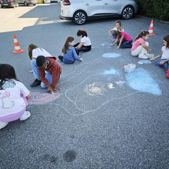 Kinder malen mit Kreide auf dem Bürgersteig, umgeben von Verkehrskegeln. Ein silberner Wagen ist in der Nähe geparkt.