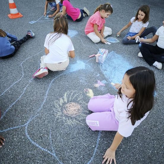 Eine Gruppe junger Mädchen sitzt auf dem Bürgersteig und malt Blumen mit Kreide. Eines der Mädchen ist in der Vorderansicht und hält ein Stück Kreide in der Hand.