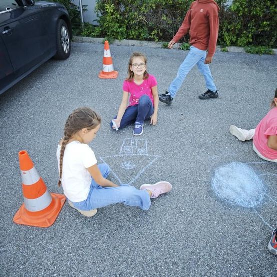 Drei junge Mädchen spielen Einkästchen auf einer Kiesauffahrt. Zwei Mädchen sitzen, während das dritte vorbeigeht. In der Nähe befinden sich zwei orangefarbene Verkehrskegel.