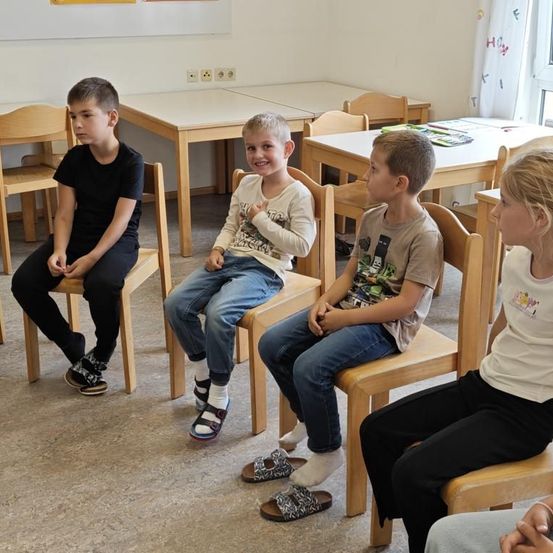 Vier Kinder sitzen in einem Klassenzimmer auf Stühlen und lächeln. Der Junge in Schwarz schaut nach unten. Der Junge in Weiß schaut in die Kamera. Der Junge in Grau schaut weg. Ein Mädchen in Weiß hört zu.