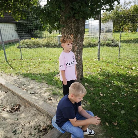 Zwei junge Jungen stehen draußen, einer sitzt auf einer Bank und der andere steht neben einem Baum mit einem rosa T-Shirt. Dahinter befindet sich ein eingezäunter Bereich mit einem Haus und Bäumen.