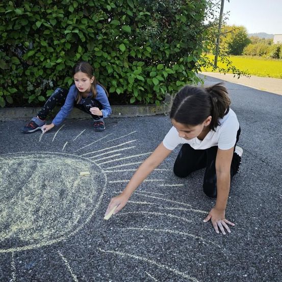 Zwei Mädchen knien auf dem Asphalt und zeichnen mit Kreide eine Sonne. Eines hält ein Stück Kreide in der Hand. Hinter ihnen steht ein großer grüner Busch.