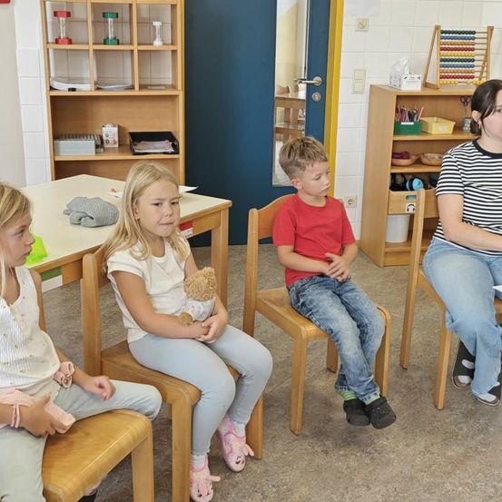 Vier Kinder sitzen auf Stühlen in einem Klassenzimmer. Ein Mädchen hält einen Teddybären und ein Junge in einem roten T-Shirt schaut weg. Eine Lehrerin sitzt neben ihnen, mit Regalen und einer blauen Tür im Hintergrund.