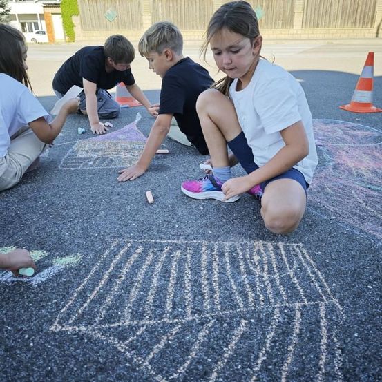 Vier Kinder zeichnen mit Kreide auf dem Asphalt. Drei sind Jungen und eines ist ein Mädchen. Sie tragen Freizeitkleidung. Der Boden ist mit Kegeln und Kreide markiert.