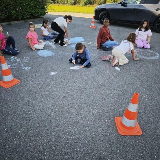 Eine Gruppe von Kindern malt mit Kreide auf dem Boden, umgeben von Verkehrskegel, mit einem geparkten Auto in der Nähe.