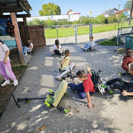 Kinder sitzen auf ihren faltbaren Rollern in einem schattigen Bereich außerhalb eines Gebäudes. Einige sind auf dem Boden, während andere sitzen. Eine Frau steht auf der linken Seite.