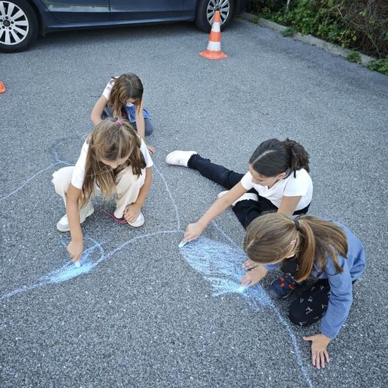Vier junge Mädchen zeichnen mit Kreide auf dem Asphalt, ein Mädchen kniet, während die anderen sitzen. Dahinter steht ein Poller.