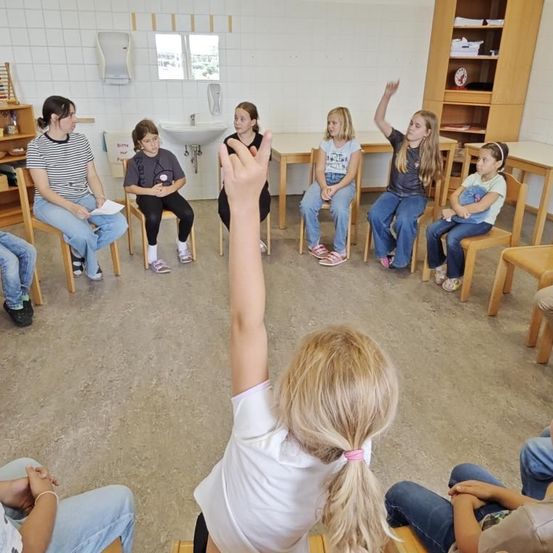 Eine Gruppe von Kindern sitzt in einem Klassenzimmer im Kreis. Ein Mädchen hebt die Hand. Hinter ihnen befinden sich Regale, ein Waschbecken und ein Fenster.