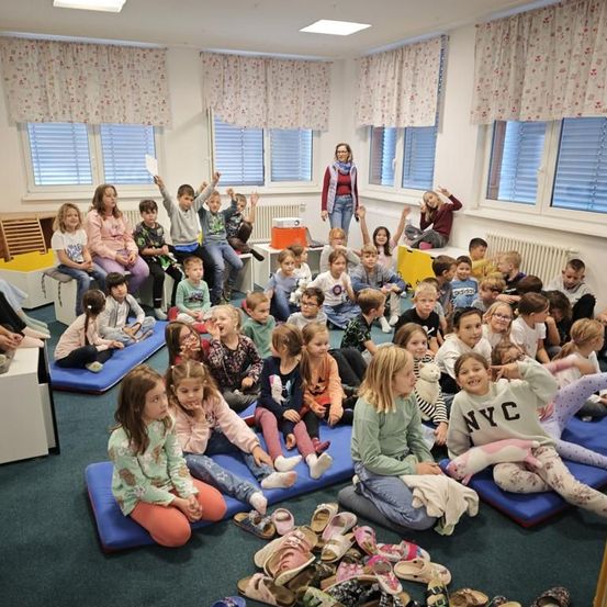 Ein Klassenzimmer voller Kinder, die auf blauen Matten sitzen, einige mit Schuhen um sie herum verstreut. Ein Erwachsener steht in der Ecke, und ein Projektor ist in der Nähe. Der Raum hat weiße Wände und Fenster mit Vorhängen.