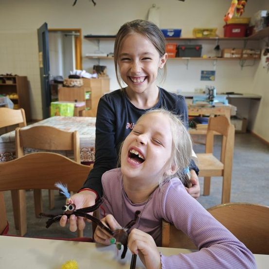 Zwei junge Mädchen lachen im Klassenzimmer und halten eine Spinnenbastelei in den Händen. Sie sind von Holzstühlen und Tischen umgeben. Dahinter befinden sich Regale mit Aufbewahrungsboxen und ein Schreibtisch mit verschiedenen Gegenständen.