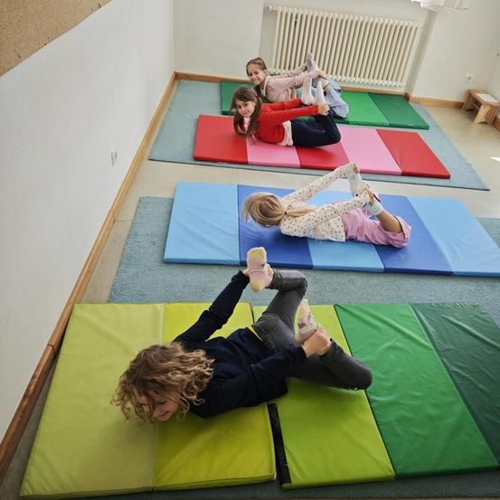 Vier junge Mädchen machen Übungen auf mehrfarbigen Matten in einem Raum. Ein Heizkörper ist an der Wand montiert.