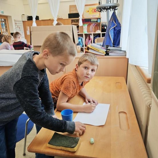Zwei Jungen in einem Klassenzimmer, einer schreibt mit einem Stift auf Papier, während der andere steht und das Papier betrachtet. Ein blauer Becher und ein grünes Objekt liegen auf dem Tisch.