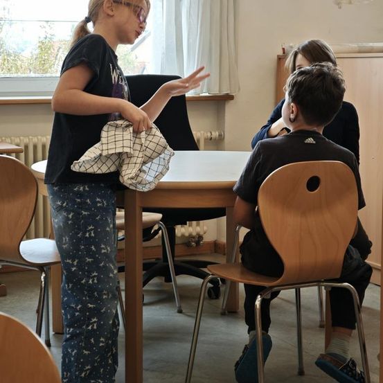 Ein Mädchen steht in der Nähe eines Tisches und hält ein Tuch in der Hand, während zwei Kinder auf Stühlen sitzen. Der Raum hat weiße Wände, ein Fenster mit einem weißen Vorhang und eine Heizung.