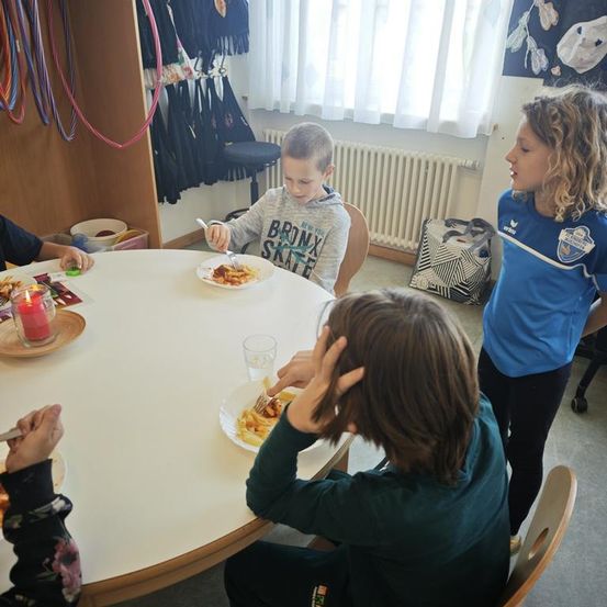 Fünf Kinder sitzen um einen Tisch herum und essen. Ein Kind hält eine Gabel in der Luft, eine Mädchen steht neben ihnen und ein Junge schaut auf sein Essen.