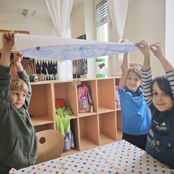 Drei junge Kinder in einem Klassenzimmer halten ein großes Blatt Papier mit Zeichnungen hoch. Ein Tisch ist mit einer Tischdecke mit Punkten bedeckt, und Regale voller Spielzeug und Rucksäcke befinden sich hinter ihnen.