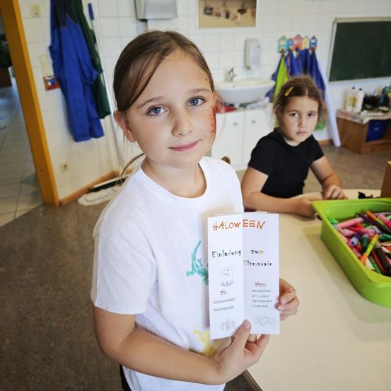 Ein junges Mädchen hält eine Halloween-Einladung in der Hand und steht in einem Klassenzimmer mit einem anderen Mädchen am Schreibtisch. Im Hintergrund befinden sich eine Tafel und eine Aufbewahrungsbox mit Buntstiften.