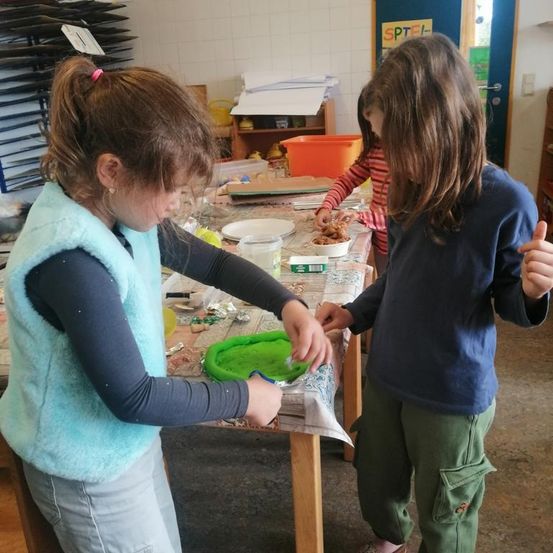 Drei Kinder arbeiten an einem Bastelprojekt an einem Tisch. Ein Kind in einer Weste arbeitet an einem grünen Gegenstand, ein anderes in einem gestreiften Shirt isst in der Nähe und das dritte gibt Daumen hoch.