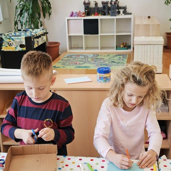 Zwei Kinder sind in einem Klassenzimmer, ein Junge schneidet ein Stück Pappe und das andere Mädchen zeichnet auf einem Blatt Papier.