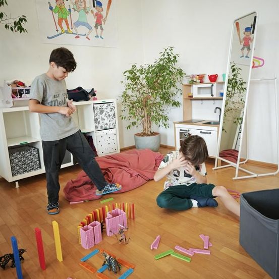 Zwei Jungen spielen mit bunten Dominosteinen auf einem Holzboden. Ein Junge sitzt und bedeckt sein Gesicht, während der andere in der Nähe steht. Ein Topf mit einer Pflanze und eine Mikrowelle befinden sich im Hintergrund.