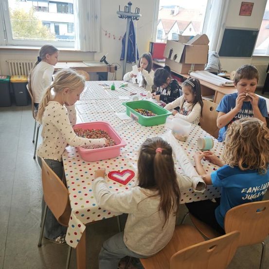 Eine Gruppe von Kindern sitzt in einem Klassenzimmer um einen Tisch herum und ist mit einer Aktivität beschäftigt. Der Tisch ist mit einem tupfigen Tuch bedeckt, und verschiedene Bastelutensilien sind darauf platziert. Ein Kind isst, während andere sich auf ihre Aufgaben konzentrieren.