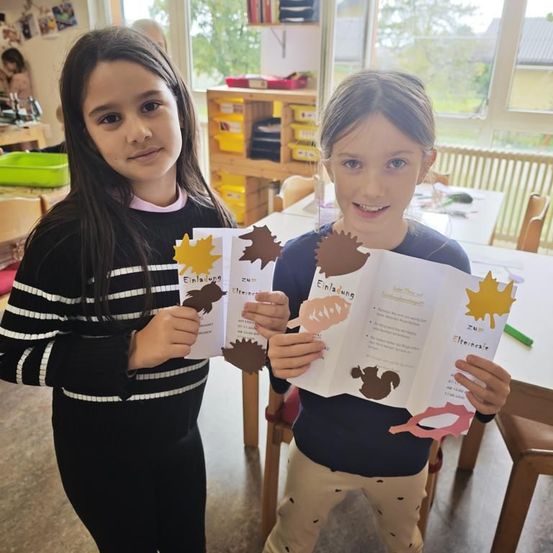 Zwei Mädchen stehen in einem Klassenzimmer und halten Herbstbasteleien in den Händen. Sie lächeln und schauen in die Kamera. Hinter ihnen ist ein Tisch mit verschiedenen Gegenständen zu sehen, und eine Frau steht in der Nähe eines Fensters.