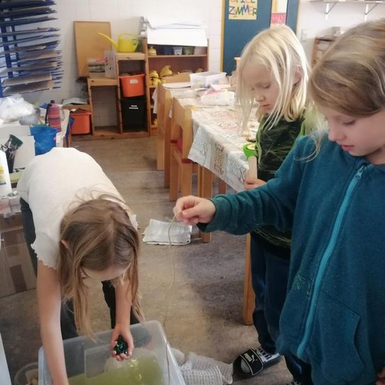 Drei Kinder sind in einem Raum mit Tischen und Stühlen und beschäftigen sich mit einer Aktivität. Ein Mädchen beugt sich über einen Behälter, während die anderen zuschauen.
