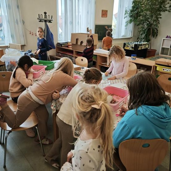 Kinder arbeiten in einem Klassenzimmer an einem Bastelprojekt. Einige Kinder sitzen an einem Tisch mit bunten Gegenständen in Schalen, während andere stehen oder auf Stühlen sitzen. Ein Mädchen steht in der Nähe einer Tafel.