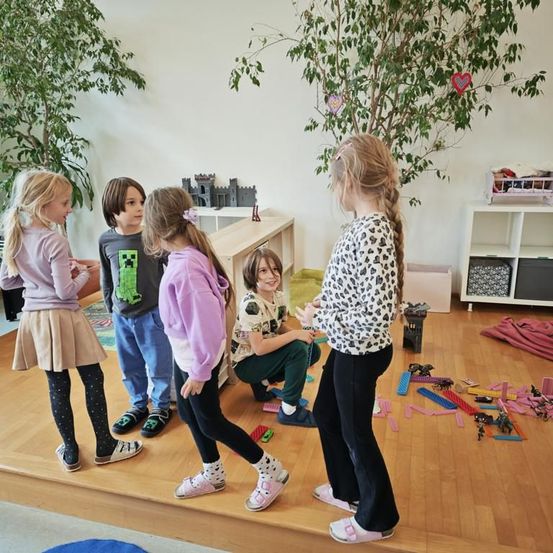 Fünf Kinder stehen in einem Raum mit Holzboden, umgeben von Spielzeug. Ein Kind mit einem Leoparden-Print-Shirt führt eine Gruppe an, während andere zusehen. Topfpflanzen und ein Regal sind im Hintergrund.