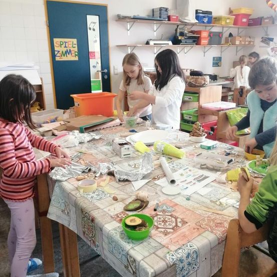Kinder beschäftigen sich in einem Klassenzimmer mit einer Kunstaktivität. Sie haben einen Tisch, der mit verschiedenen Kunstmaterialien bedeckt ist. Zwei Mädchen in weißen Shirts greifen nach Materialien.