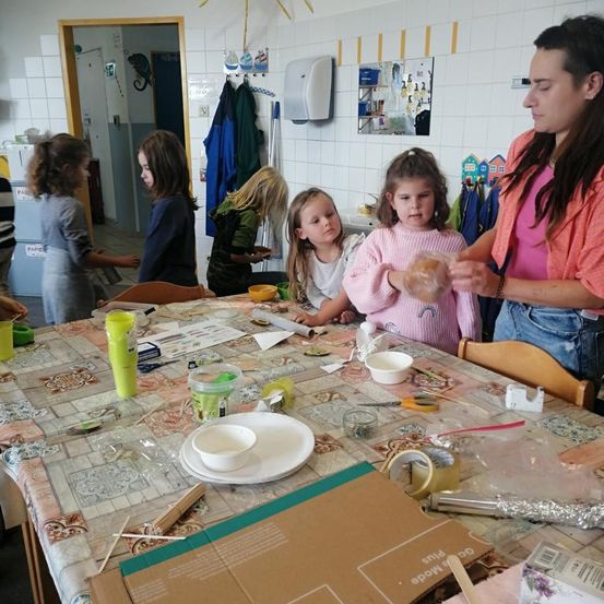 Eine Frau unterrichtet eine Gruppe von Mädchen bei einem Bastelprojekt an einem Tisch, umgeben von verschiedenen Materialien und Werkzeugen.