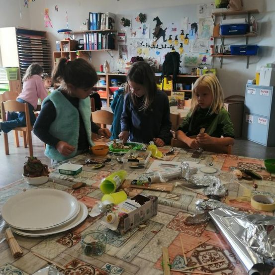 In einem Klassenzimmer arbeiten vier Mädchen an einem Tisch mit Tellern, Schüsseln und Bastelutensilien. Sie scheinen zu basteln. Der Raum hat Regale mit Büchern, Papieren und einem Schrank.