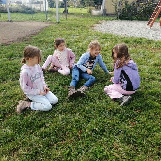 Vier junge Mädchen sitzen auf einem Grasfeld, möglicherweise in einem Spielplatz, wobei ein Mädchen lächelt. Sie tragen Jacken und Turnschuhe. Dahinter befindet sich ein Metallzaun mit einer Holzleiter.