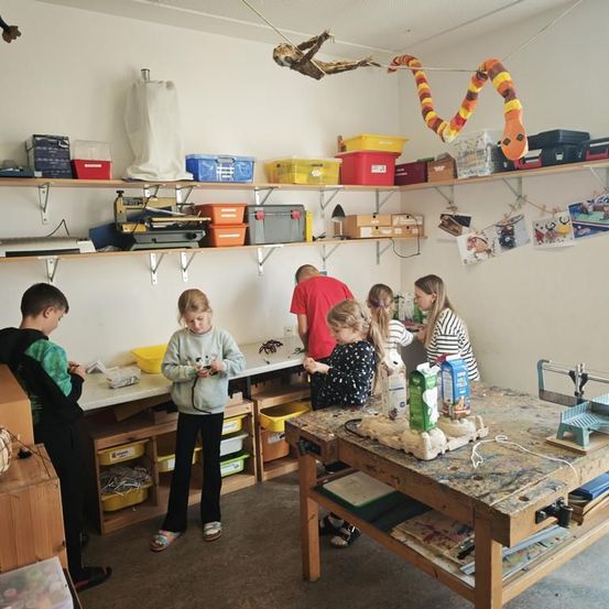 Eine Gruppe von Kindern arbeitet in einer Werkstatt, wobei ein Mann und eine Frau die Aufsicht führen. Verschiedene Werkzeuge und Materialien sind auf den Tischen und Regalen.