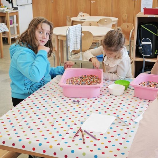 Zwei junge Mädchen sitzen an einem Tisch mit farbenfrohen Streuseln in rosa Behältern und tragen Freizeitkleidung. Ein Mädchen hat eine Brille, und sie scheinen sich auf ihre Aktivität zu konzentrieren. Es gibt Schüsseln, Papier und Stifte auf dem Tisch, und Regale mit Gegenständen im Hintergrund.